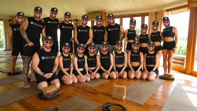A group of people in matching black "strcUSX" uniforms and red sunglasses pose together in a yoga-like setting, with one man seated prominently in front.

The image relates to the post by showing the strcUSX community/team in a humorous, unified pose—possibly reflecting the discipline and consistency ("mint k$ đều đều") praised in the caption about @Manners_ST.
