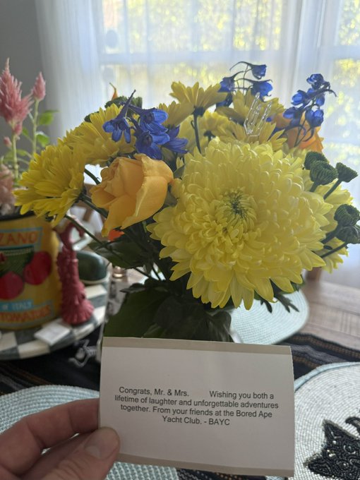 A hand holding a congratulatory card that reads 'Congrats, Mr. & Mrs. Wishing you both a lifetime of laughter and unforgettable adventures together. From your friends at the Bored Ape Yacht Club. - BAYC,' with a vase of yellow and blue flowers in the background.