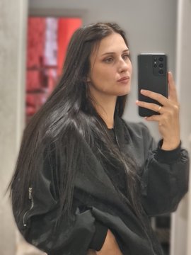 A woman with long dark hair wearing a black leather jacket takes a mirror selfie with her smartphone.