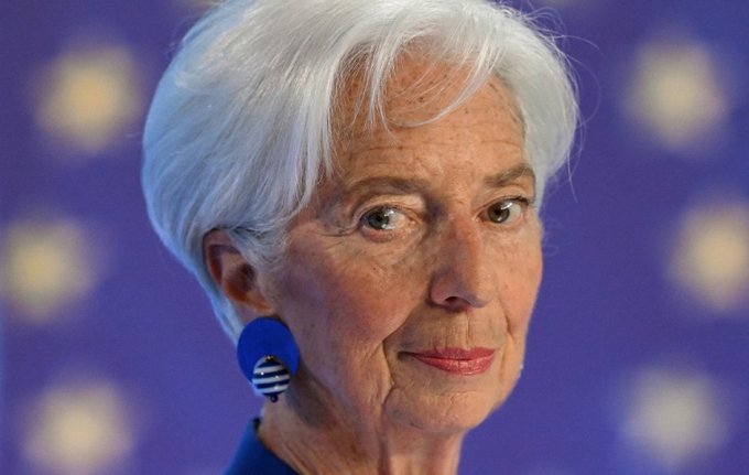 The photo features Christine Lagarde, the President of the European Central Bank, shown in a close-up portrait with short white hair, blue circular earrings, and a serious expression against a blurred purple background. The image accompanies news about her reportedly intervening to block Binance from obtaining a MiCA license in Greece.