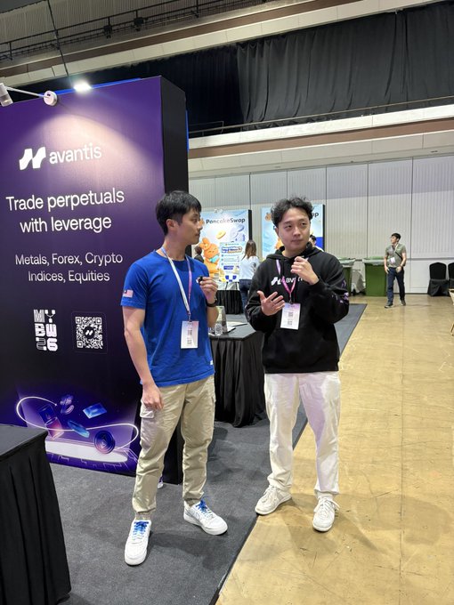 Two men standing at a trading convention booth with an 'avantis' banner advertising 'Trade perpetuals with leverage' for Metals, Forex, Crypto, Indices, and Equities, featuring an MEXC logo and QR code.