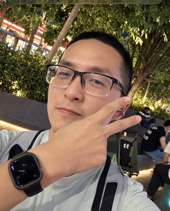 A selfie shows a young man wearing black-rimmed glasses, a light blue t-shirt, and an Apple Watch, posing outdoors at night with trees and seating areas in the background.
