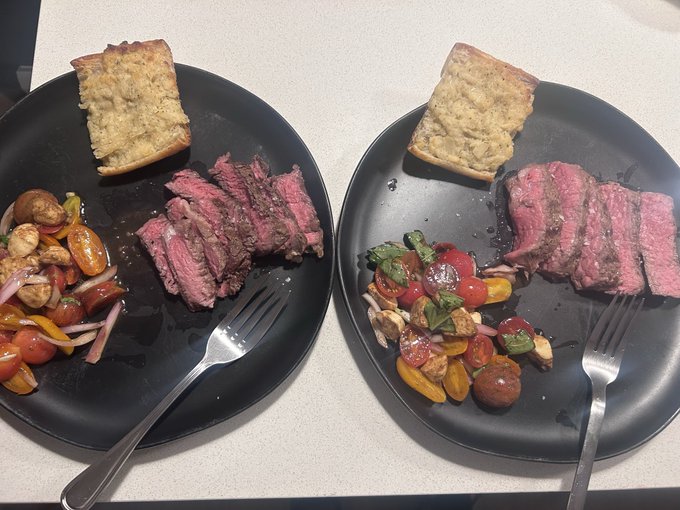 Two black plates hold sliced steak, cheesy bread, and a salad, each with a fork resting on the plate.