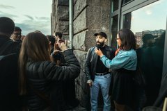 A man in a black jacket is being interviewed by reporters and photographers with cameras outside a building.