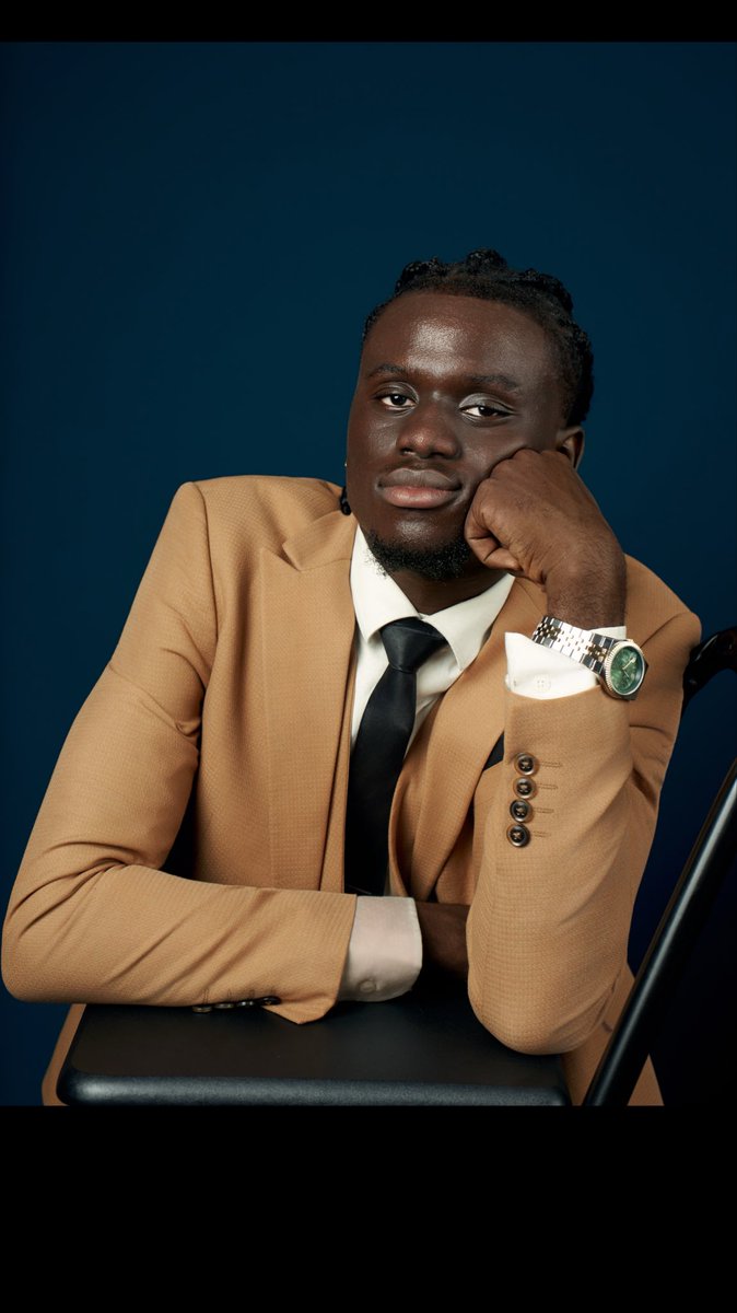 A portrait of a man in a tan suit and black tie resting his chin on his hand, wearing a silver watch, against a dark blue background.