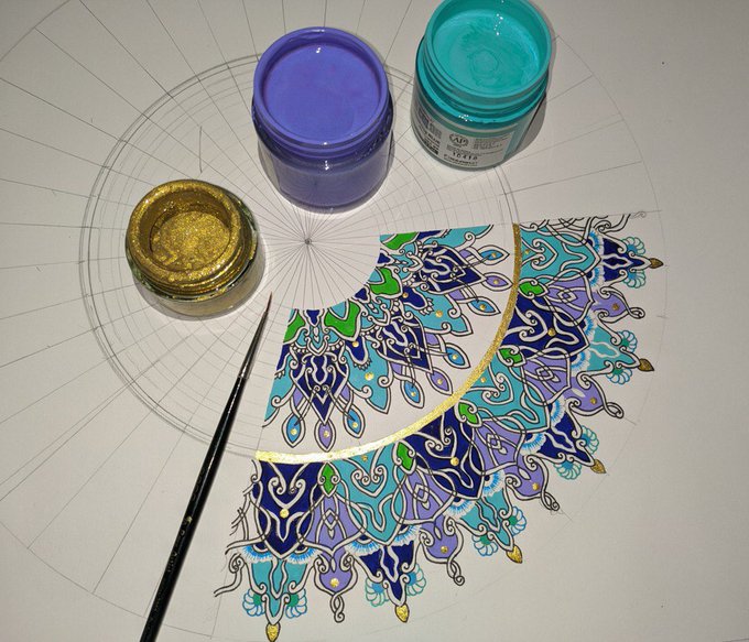 A flat-lay of an intricate mandala-style artwork in progress with blues, purples, and greens, accompanied by jars of purple and teal paint, gold pigment, a brush, and pencil guidelines.