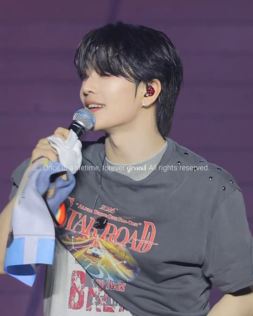 The image features a young man with short hair wearing a black t-shirt, holding a microphone while smiling. There are descriptions of other aspects such as his appearance, his shirt, and even some text that references a book or a series named Starroad. The overall atmosphere of the photo seems positive and upbeat, reflecting the cheerful demeanor of the young man captured in it.
