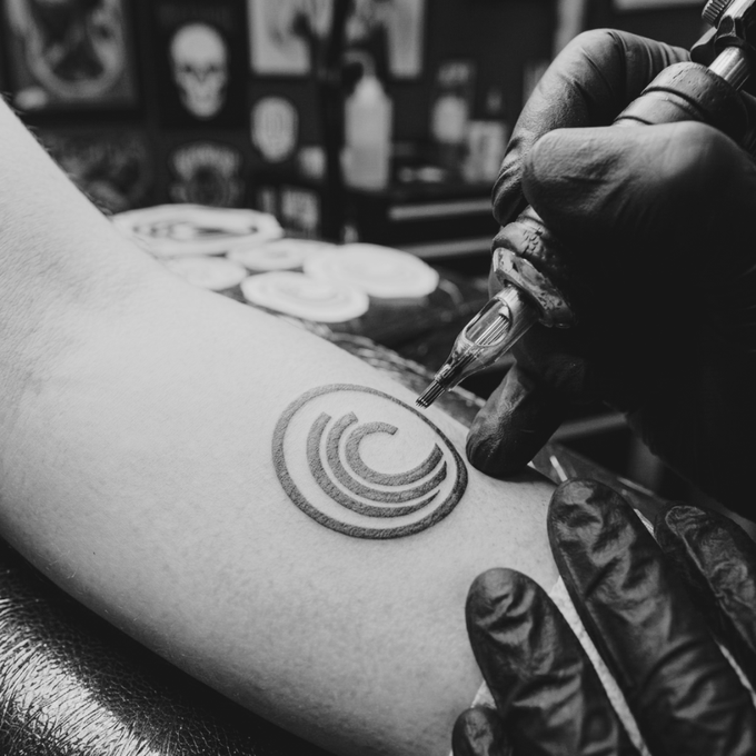 A black and white close-up of gloved hands using a tattoo machine to ink a circular spiral design onto someone's arm.