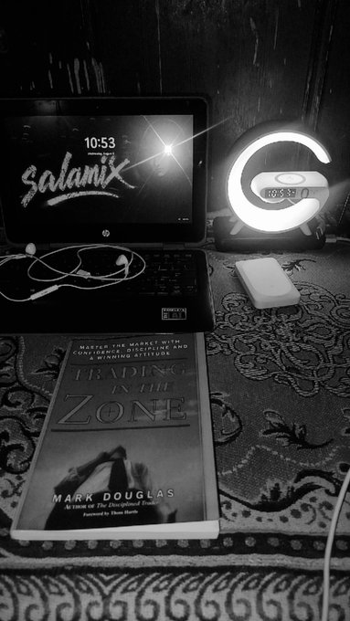 A black-and-white photo of a workspace shows an HP laptop displaying 'Salamix' at 10:53, a glowing Logitech G clock showing 10:53, earphones, a mouse, and Mark Douglas's book 'Trading in the Zone' on a patterned rug.