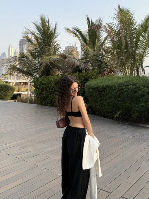 A woman with curly hair wearing a black outfit, holding a white jacket, standing on a wooden boardwalk with palm trees and a city skyline in the background.
