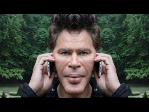 A man holds two cell phones to his ears with a serious expression, set against a forest background.