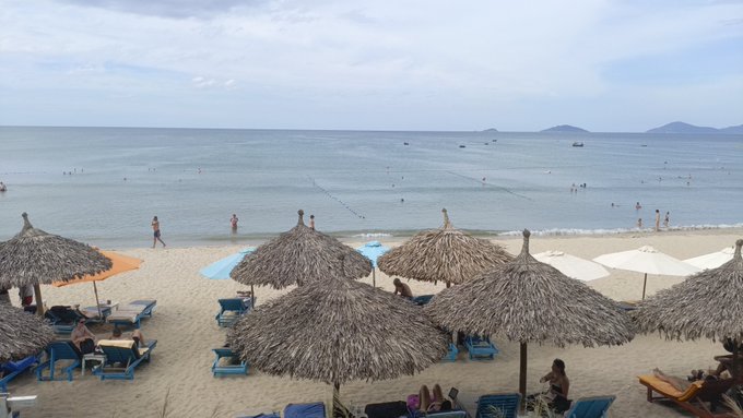 An elevated view of a sandy beach with thatched umbrellas, lounge chairs, people swimming in calm ocean water, and distant mountains on the horizon.