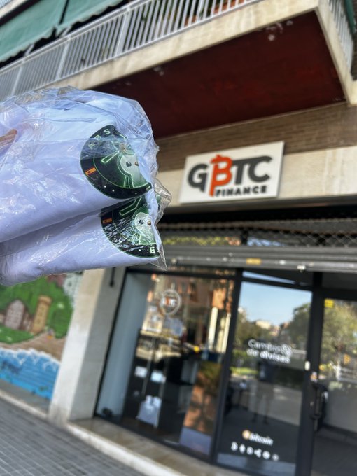 A person holds plastic-wrapped merchandise featuring circuit board graphics outside a storefront bearing a 'GBTC Finance' sign.