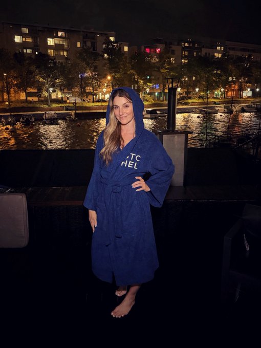 A smiling woman stands outdoors at night wearing a blue hooded robe with white lettering, with a lit waterfront, boats, and city buildings visible in the background.