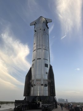 A large silver Starship rocket standing vertically on a launch pad under a cloudy sky.