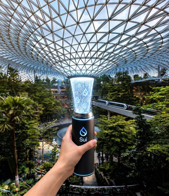A hand holds a black cylindrical container labeled "Sui" with water spraying upward, set against the backdrop of Jewel Changi Airport's indoor waterfall and glass dome.