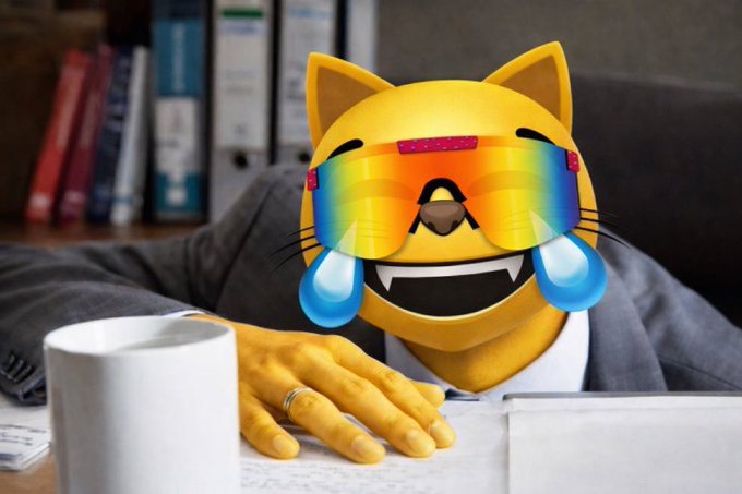 A cat emoji wearing rainbow sunglasses with tears of joy sits at a desk with a coffee mug and papers.