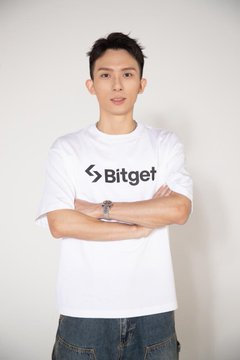 A man wearing a white t-shirt with the Bitget logo and text standing with his arms crossed against a plain white background.