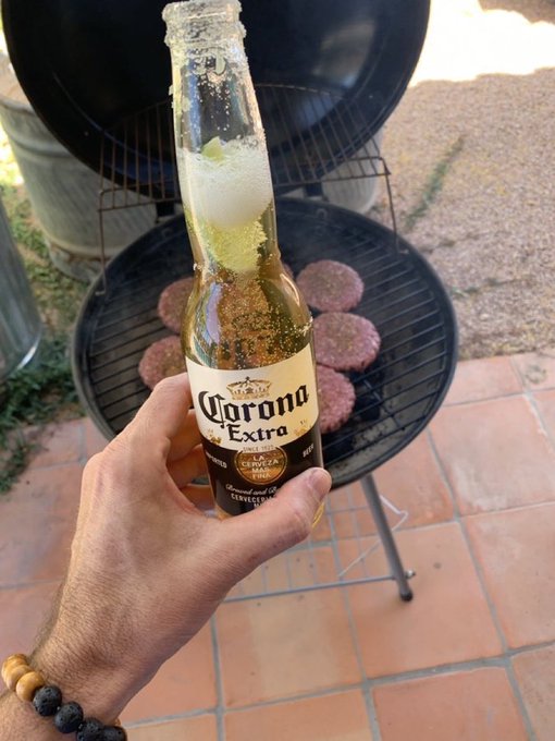 A hand holds a chilled Corona Extra beer bottle in front of a round charcoal grill cooking several raw burger patties.