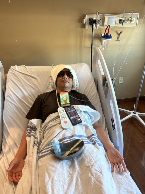 A man lies in a hospital bed with a head wrap, sunglasses, and a remote control on his chest, with medical equipment visible on the wall behind him.