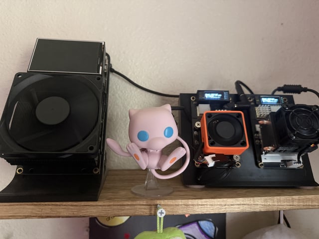 A pink Mew Pokémon figurine stands on a wooden shelf between an open black gaming console with exposed cooling fans and internal components.
