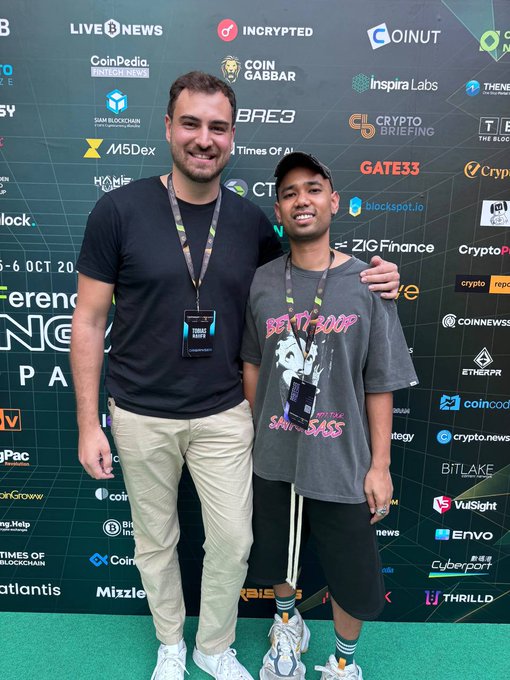 Two smiling men pose together at a crypto/blockchain conference, standing in front of a step-and-repeat banner featuring logos like LIVE NEWS, INCRYPTED, COINUT, M5Dex, ZIG Finance, and ETHERAX.