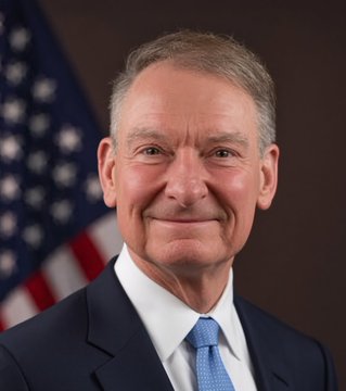 An official portrait of a gray-haired man in a dark suit and blue tie, smiling in front of an American flag.