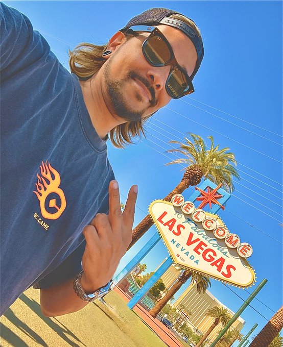A selfie of a man with long hair, sunglasses, and a black cap making a peace sign, wearing a dark blue t-shirt with an orange BC.GAME flame logo, posing in front of the 'Welcome to Fabulous Las Vegas Nevada' sign.