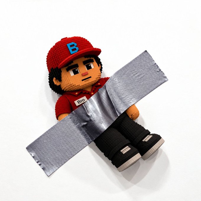 A brick-built figure of a boy wearing a red cap with a blue 'B', a red shirt with a 'Bro' nametag, and black pants, lying on his back while taped to a white background in an X shape.