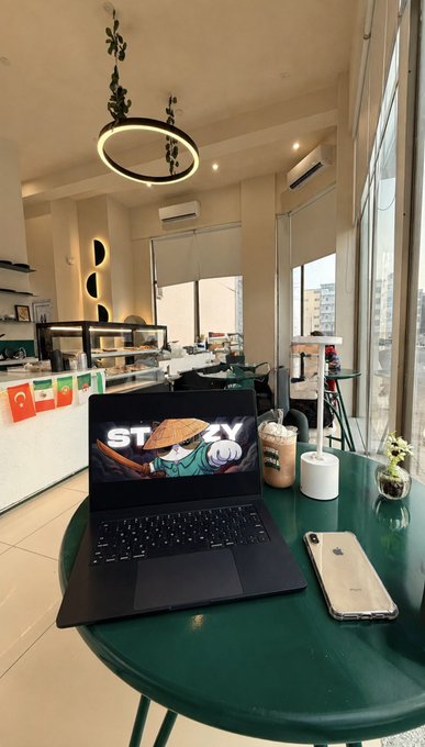 A workspace scene showing a laptop with a 'STAY' anime wallpaper, a phone, and an iced coffee on a round green table, with a café interior, hanging ring pendant light, air conditioner, and national flags visible in the background.