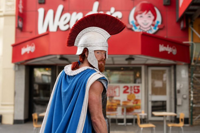 A man dressed in a Roman centurion costume with a red-plumed helmet and blue cape standing in front of a Wendy's restaurant with a "$1 $5" promotional sign.