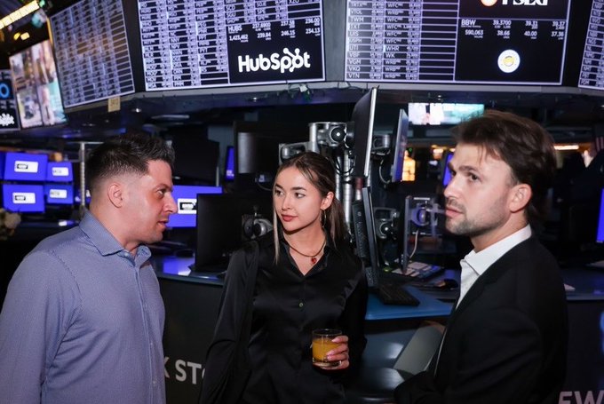 Three people conversing on what appears to be the floor of the New York Stock Exchange, with HubSpot signage and stock tickers visible in the background.