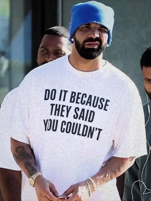 Drake wearing a blue beanie and a white t-shirt that reads 'DO IT BECAUSE THEY SAID YOU COULDN'T,' walking outdoors with others.