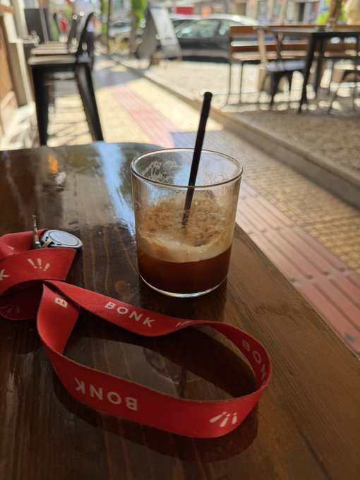 A half-finished iced coffee in a glass with a black straw sits on a wooden table next to a red lanyard reading 'BONK', with a patio and parked cars visible in the background.