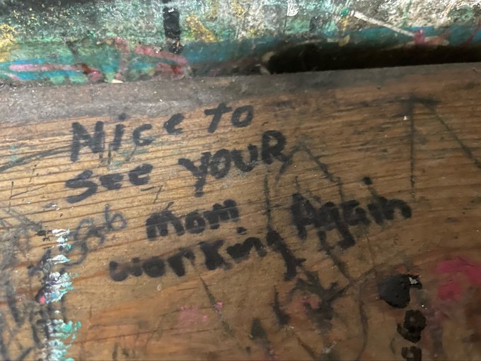 A weathered wooden surface with handwritten graffiti reading 'Nice to See your mom! Again' along with other partially legible scribbles.