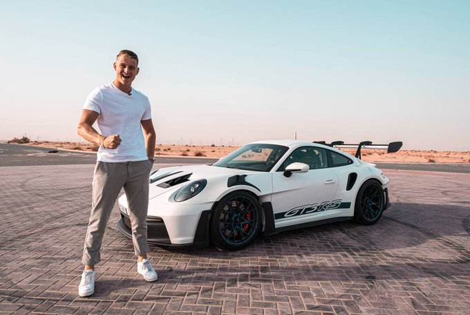 The image features a man standing next to a white sports car, possibly a Porsche 911 GT3 Spyder, parked on a brick road or pavement. The man appears to be posing for a photo with the car as his backdrop. The caption of the image includes some text that reads "CRYPTO ROVER WILL LONG THE BOTTOM AND MAKE MILLIONS NEXT BULL RUN!" This phrase is likely an inside joke or reference to the car's capabilities, such as its speed and performance.
