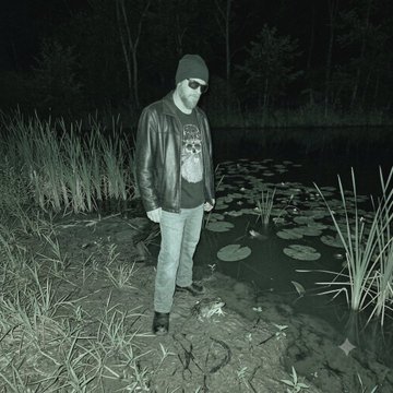 A black-and-white nighttime photograph depicts a bearded man wearing sunglasses, a beanie, and a leather jacket standing on rocks beside a pond with lily pads.