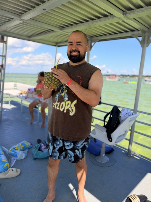A smiling bald man wearing a "Hotlops" t-shirt and patterned swim trunks holds a fresh pineapple drink on a boat over turquoise water.