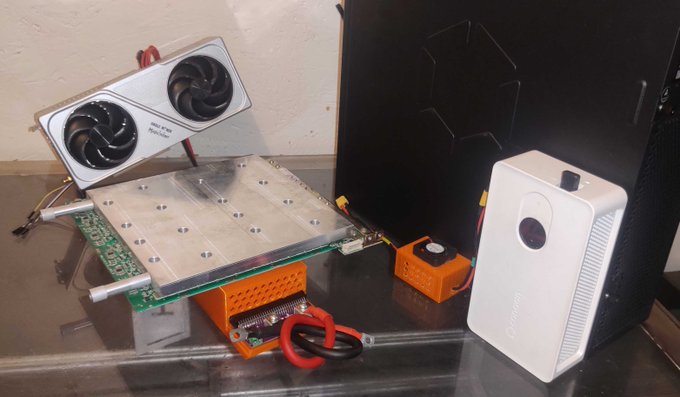 A collection of cryptocurrency mining hardware and accessories on a glass shelf, including a cooling fan assembly, a circuit board with an orange 3D-printed mount, a small orange device, a black computer case, and a white 'CoolBitX' wallet.