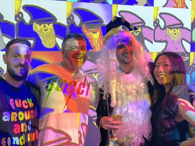 Four people pose together at what appears to be a party or event, with one person in the center wearing a white wig and holding a drink, while colorful animated projections of cartoon characters light up the wall behind them. The person on the left wears a shirt reading "fuck around and find out," and the festive, playful atmosphere with vibrant lighting reflects the post's message about Bitcoin figures like Bitboy, Eric, and Udi bringing fun back to the cryptocurrency community.