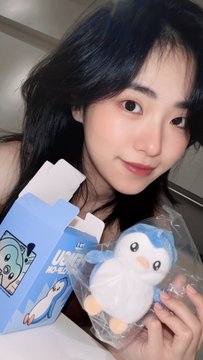 A young woman smiling as she holds a plush penguin toy and a blue box, possibly a merch purchase.
