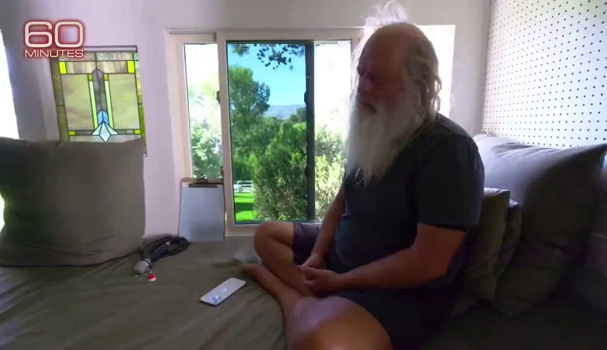 The image shows an older man sitting on a bed with his legs crossed, wearing shorts and a gray shirt. He has white hair and appears to be looking down at something or someone. The background of the image features a window overlooking trees outside. There is also a remote control placed next to him on the bed.