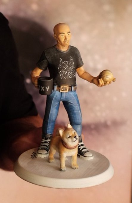 A figurine of a bald man in a black t-shirt and jeans holding a mug (marked 'XY') and a cookie, standing with a smiling Shiba Inu dog at his feet.