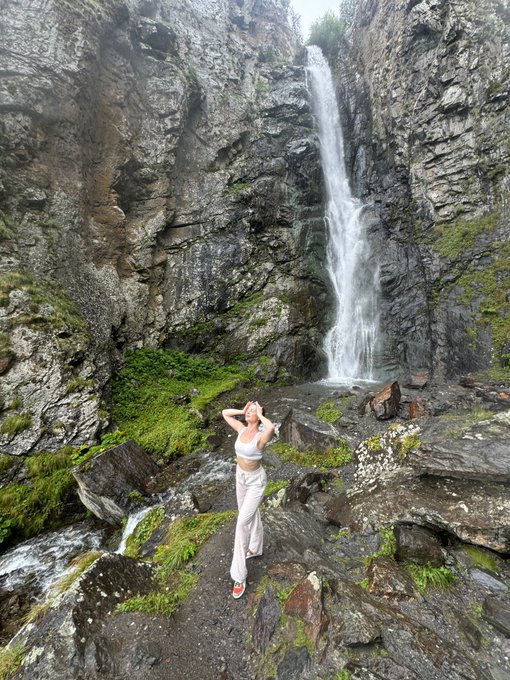 A young woman in a white top and pants poses in front of a tall waterfall cascading down rocky cliffs into a mossy gorge.