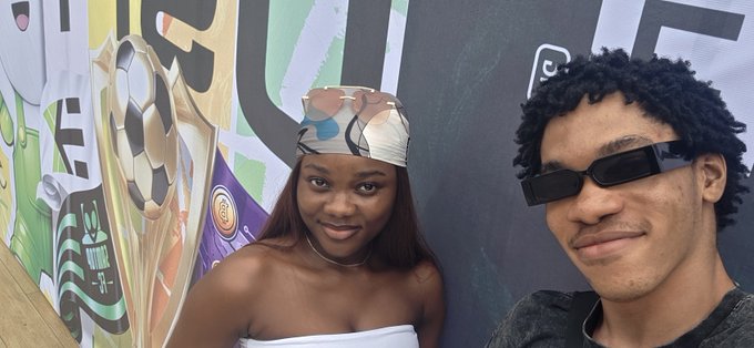 A young woman wearing a patterned headscarf and a strapless top stands next to a young man wearing black rectangular sunglasses and a dark shirt, posing in front of a colorful mural featuring a soccer trophy.