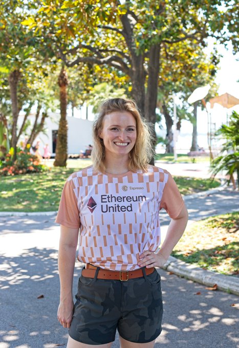 A smiling woman with blonde hair poses outdoors on a sunny day, wearing an 'Ethereum United' jersey with an 'Espresso' sponsor logo, paired with camouflage shorts.