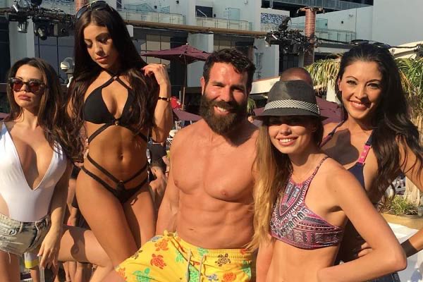 A group of five young adults pose for a photo at an outdoor pool party; a shirtless, muscular man with a beard in colorful swim trunks stands in the center, flanked by four women in bikinis.