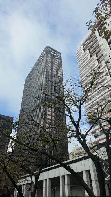A low-angle photograph of a tall modern skyscraper with circular logos near its top, framed by tree branches against a bright blue sky with scattered clouds.