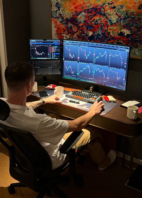 A trader sitting at a multi-monitor workstation with trading charts, using a mouse and keyboard, with mugs and a colorful abstract painting on the wall behind.