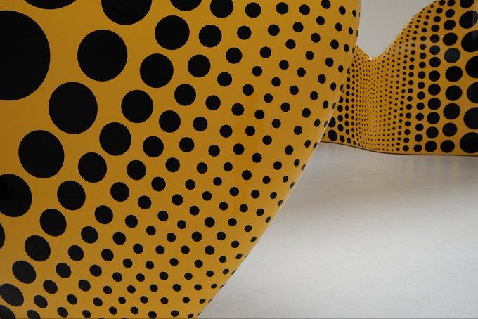 A large yellow sculpture covered in black polka dots of varying sizes is displayed in a white gallery space, characteristic of Yayoi Kusama's signature style.